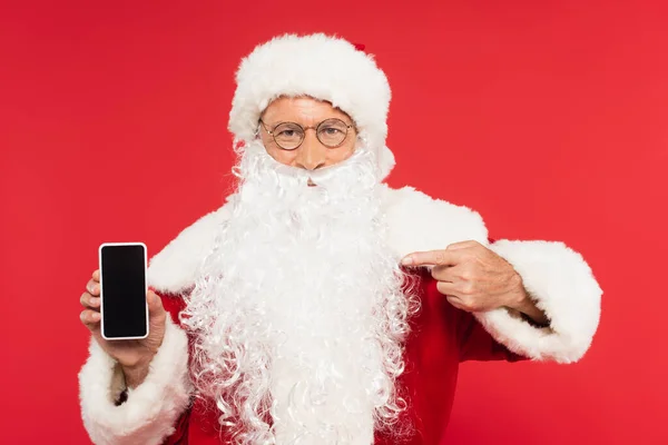 Santa Claus apuntando a teléfono inteligente con pantalla en blanco aislado en rojo - foto de stock
