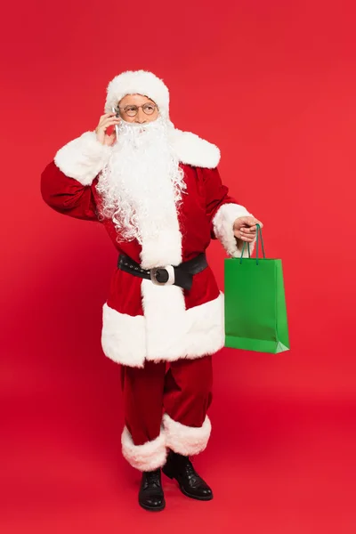 Papai Noel falando no smartphone e segurando saco de compras no fundo vermelho — Fotografia de Stock