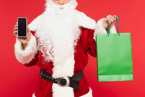 Обрезанный вид Санта-Клауса, держащего смартфон и сумку, изолированную на красном — стоковое фото