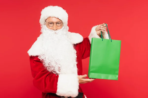 Santa claus looking at camera and holding shopping bag isolated on red — Stock Photo