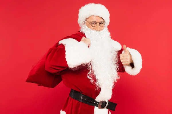 Papai Noel em traje segurando saco e mostrando como isolado no vermelho — Fotografia de Stock