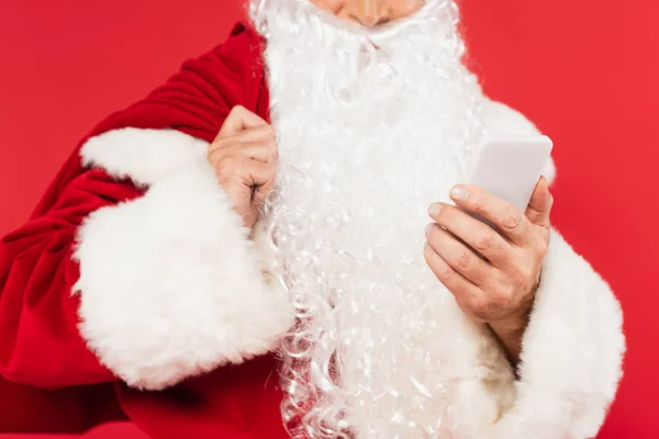 Vista cortada do smartphone na mão de Papai Noel desfocado com saco isolado no vermelho — Fotografia de Stock