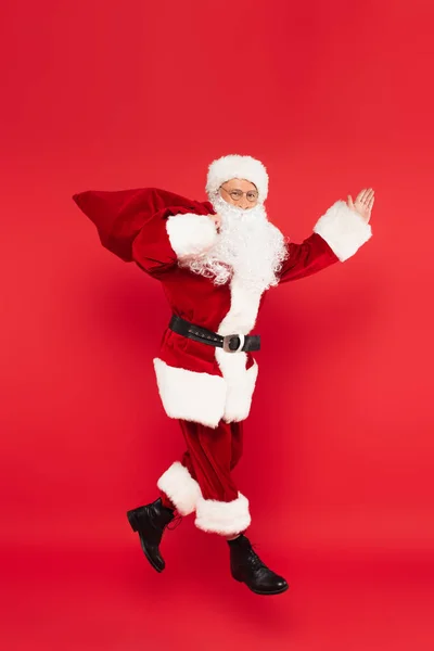 Santa claus with sack pointing with hand and levitating isolated on red — Stock Photo