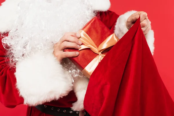 Vista recortada de Santa Claus poniendo regalo en saco aislado en rojo - foto de stock