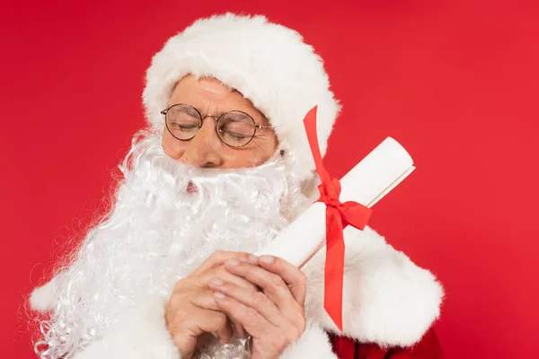 Santa claus with closed eyes holding paper with ribbon isolated on red — Stock Photo