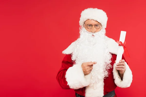 Santa claus pointing at paper with bow isolated on red — Stock Photo