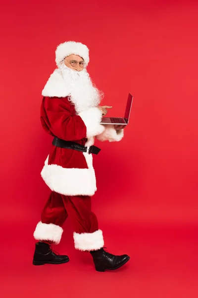 Санта Клаус указывает на ноутбук с чистым экраном на красном фоне — стоковое фото