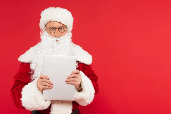 Санта-Клаус в костюмах і окулярах тримає ноутбук ізольовано на червоному — стокове фото