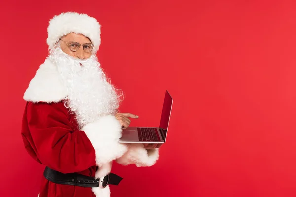 Weihnachtsmann im Kostüm zeigt auf Laptop mit leerem Bildschirm auf rotem Hintergrund — Stockfoto