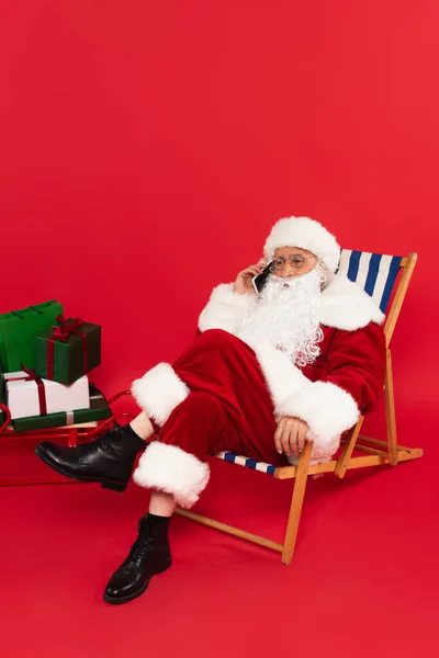 Santa Claus hablando en el teléfono inteligente en la silla de cubierta cerca del trineo con regalos sobre fondo rojo - foto de stock