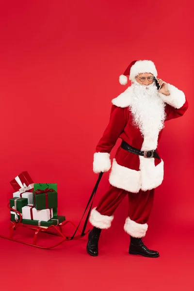 Santa hablando en smartphone y tirando del trineo con regalos sobre fondo rojo - foto de stock