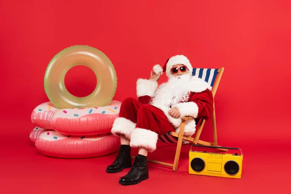 Santa Claus en gafas de sol y disfraz sentado en la silla de cubierta cerca de los anillos de natación y boombox sobre fondo rojo - foto de stock