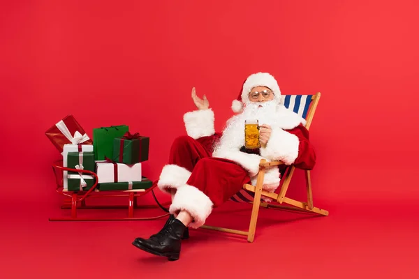 Santa Claus sosteniendo vaso de cerveza en la silla de cubierta cerca del trineo con regalos sobre fondo rojo - foto de stock