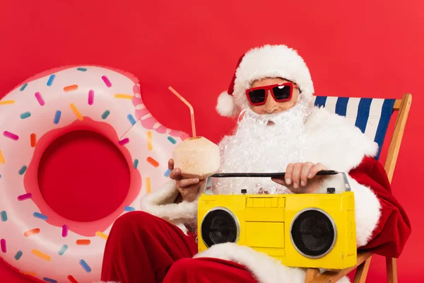 Santa dans les lunettes de soleil tenant cocktail dans la noix de coco et boombox sur chaise longue près de l'anneau de natation sur fond rouge — Photo de stock