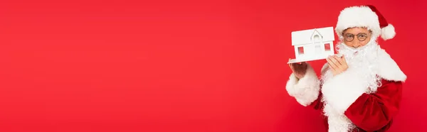 Santa claus dans des lunettes tenant modèle maison isolé sur rouge, bannière — Photo de stock