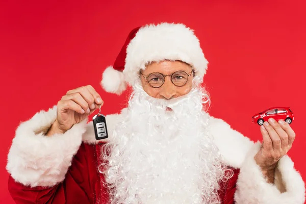 Weihnachtsmann mit Brille hält Automodell und Schlüssel isoliert auf Rot — Stockfoto