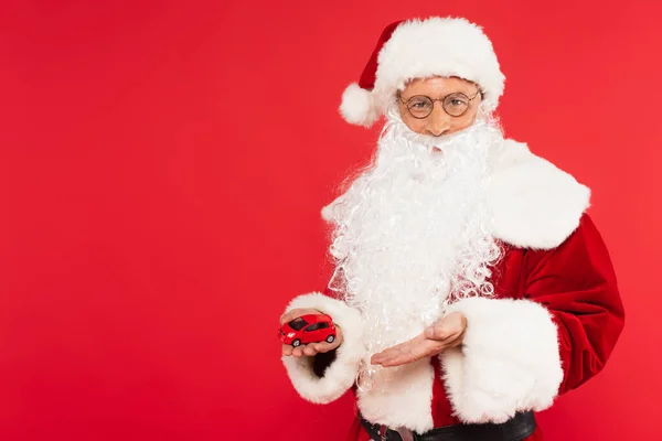 Santa in hat and eyeglasses pointing at model of car isolated on red — Stock Photo