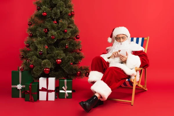 Santa Claus con teléfono inteligente en la silla de cubierta cerca del árbol de Navidad y regalos sobre fondo rojo - foto de stock