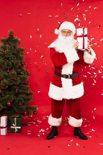 Santa claus holding present near christmas tree and confetti on red background — Stock Photo