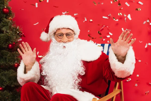 Санта-Клаус махає руками на стільці біля ялинки та конфетті на червоному тлі — стокове фото