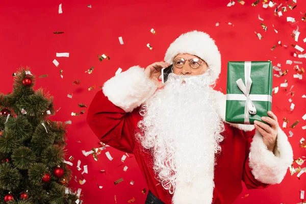 Santa claus talking on smartphone and holding gift under confetti near christmas tree on red background — Stock Photo