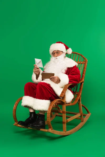 Weihnachtsmann hält Briefumschläge auf Schaukelstuhl auf grünem Hintergrund — Stockfoto