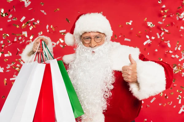 Papai Noel mostrando como e segurando sacos de compras sob confete no fundo vermelho — Fotografia de Stock