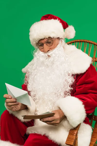 Weihnachtsmann mit Brille hält Briefumschläge auf Korbstuhl isoliert auf Grün — Stockfoto