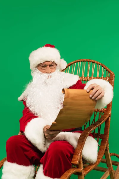 Weihnachtsmann in Kostüm und Brille hält Papier auf Korbstuhl isoliert auf Grün — Stockfoto