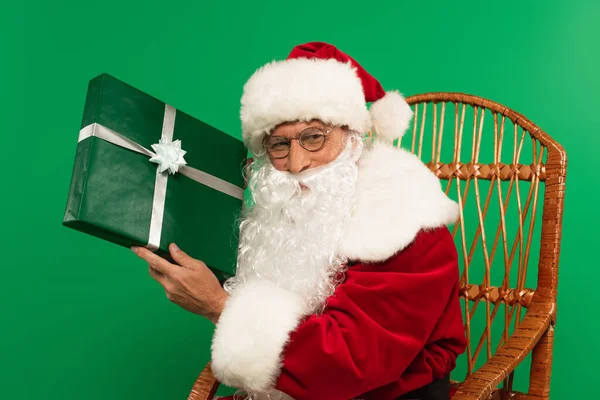 Santa claus in eyeglasses holding present on wicker chair isolated on green — Stock Photo