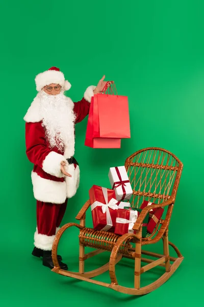 Weihnachtsmann im Kostüm hält Einkaufstüten neben Geschenken auf Korbsessel auf grünem Hintergrund — Stockfoto