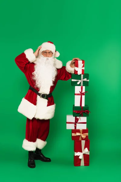 Worried santa claus standing near stacked presents on green background — Stock Photo