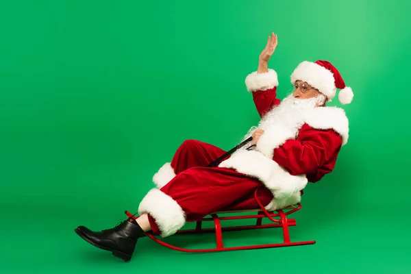Санта-Клаус махає рукою, сидячи на санях на зеленому фоні — стокове фото