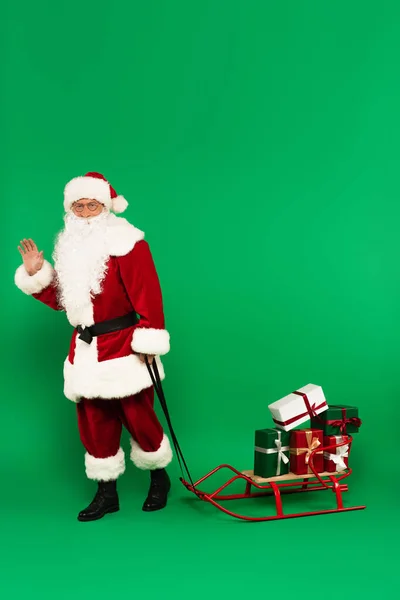 Santa claus waving hand at camera near presents on sleigh on green background — Stock Photo