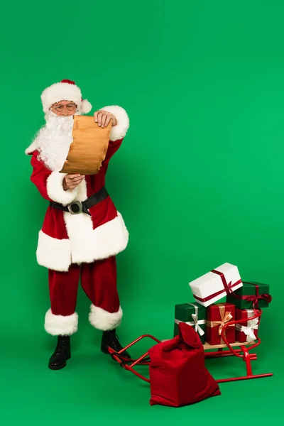 Weihnachtsmann hält Papier in der Nähe von Geschenken auf Schlitten und Sack auf grünem Hintergrund — Stockfoto