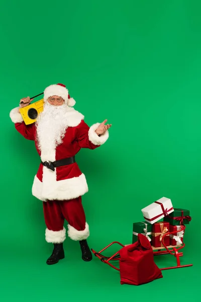 Papai Noel com boombox de pé perto de presentes no trenó e saco no fundo verde — Fotografia de Stock