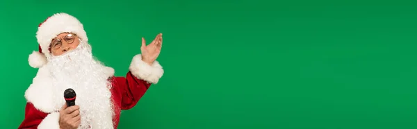 Santa Claus en traje sosteniendo micrófono y apuntando con la mano aislada en verde, bandera - foto de stock