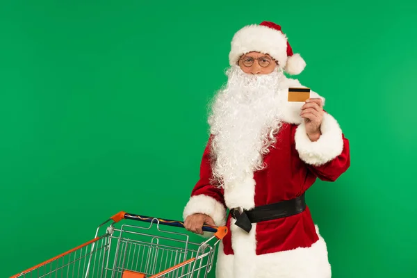 Santa claus holding credit card near shopping cart isolated on green — Stock Photo