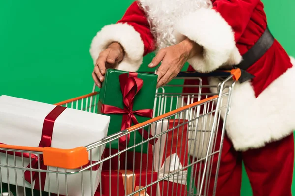 Vista recortada de Santa Claus borrosa poniendo presente en el carrito de la compra aislado en verde - foto de stock