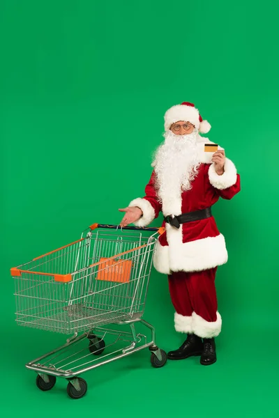 Santa claus holding credit card and pointing at shopping cart on green background — Stock Photo