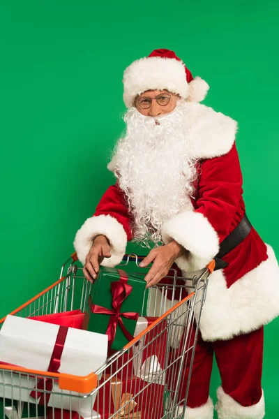 Babbo Natale in costume mettere regalo nel carrello della spesa isolato su verde — Foto stock