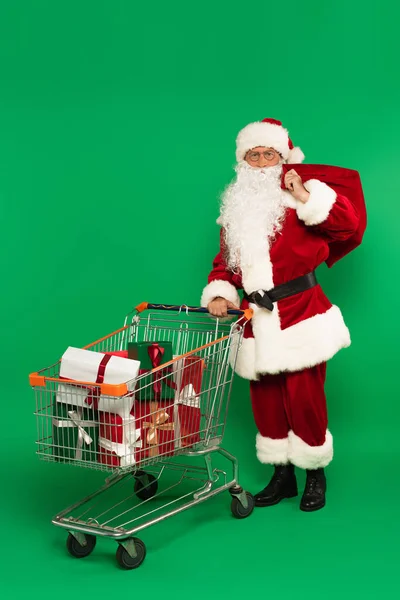 Santa Claus con saco de pie cerca del carrito de compras con regalos sobre fondo verde - foto de stock