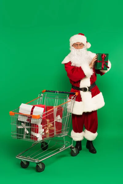 Santa Claus sosteniendo presente cerca del carrito de la compra con regalos sobre fondo verde - foto de stock
