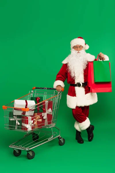 Père Noël dans des lunettes tenant des sacs à provisions près du chariot avec des cadeaux sur fond vert — Photo de stock