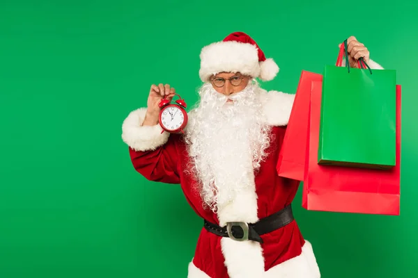 Papai Noel em traje segurando despertador e sacos de compras isolados em verde — Fotografia de Stock