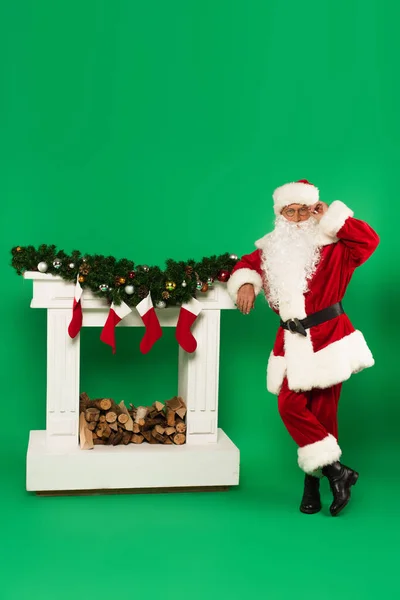 Santa claus adjusting eyeglasses near fireplace with christmas stockings on green background — Stock Photo