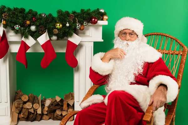 Santa claus sitting on rocking chair near fireplace with christmas stockings on green background — Stock Photo