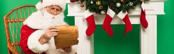 Santa claus holding paper on rocking chair near fireplace with christmas stockings on green background, banner — Stock Photo