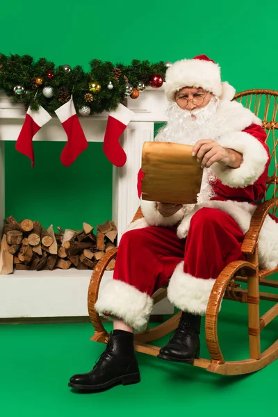 Père Noël avec du papier assis sur un fauteuil à bascule près de la cheminée avec des branches de sapin sur fond vert — Photo de stock