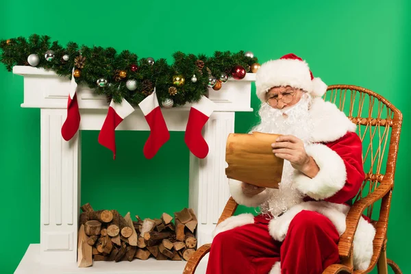 Santa claus in eyeglasses holding paper on rocking chair near decorated fireplace on green background — Stock Photo